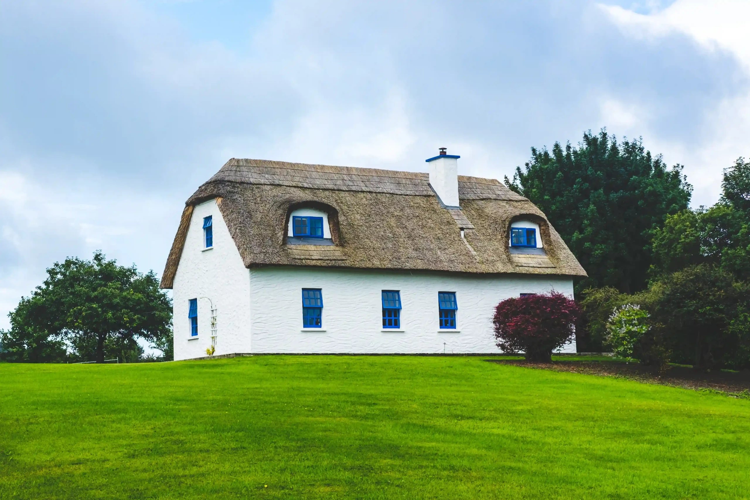 white house with a thatched roof sits nestled amidst a lush green field.